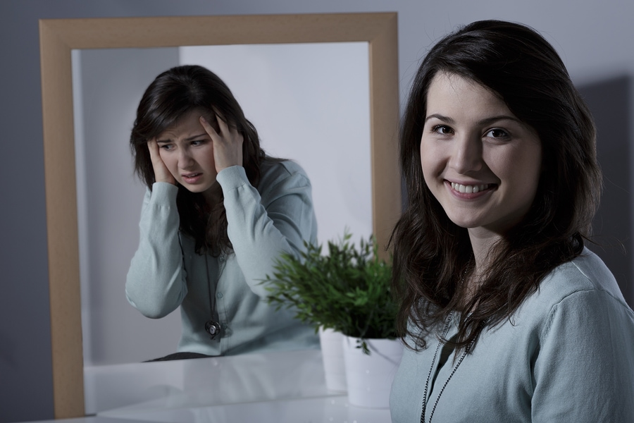 A young woman smiles in the foreground as her mirror image appears depressed or anxious.
